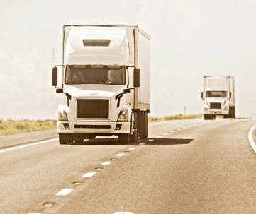 Two semi-trailer trucks are actively driving along a clear, open highway with dashed lane markings and flat surroundings.