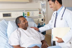 A doctor is standing beside a patient lying on a hospital bed, holding a clipboard and placing a hand on the patient's shoulder in a comforting manner