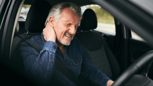 a man is driving a car, visibly uncomfortable with one hand on his neck.