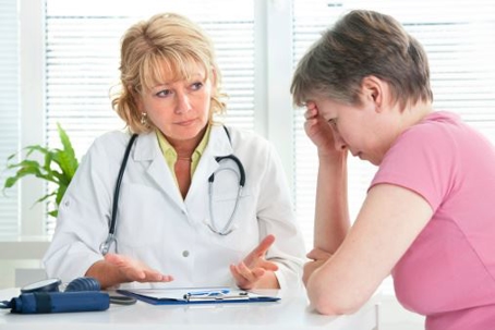 A doctor discusses health-related matters with a patient, using a clipboard for reference. A blood pressure monitor is on the desk.