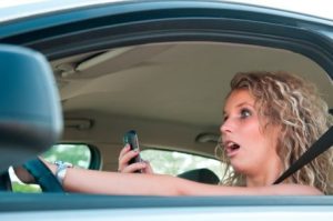 A person is sitting in the driver's seat of a car, holding a mobile phone in one hand while steering with the other.