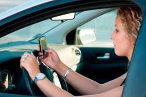 A person is seated in the driver's seat of a car, holding a smartphone with both hands while driving.