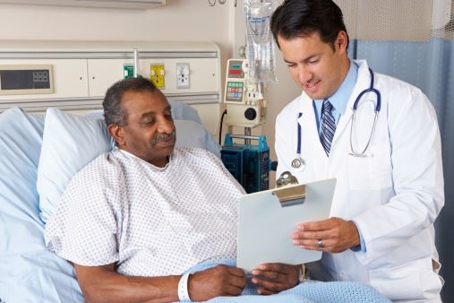 A doctor stands beside a patient lying in a hospital bed, holding a clipboard.