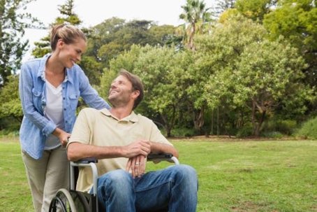 A person in a wheelchair being assisted by a caregiver in a park surrounded by trees and greenery.