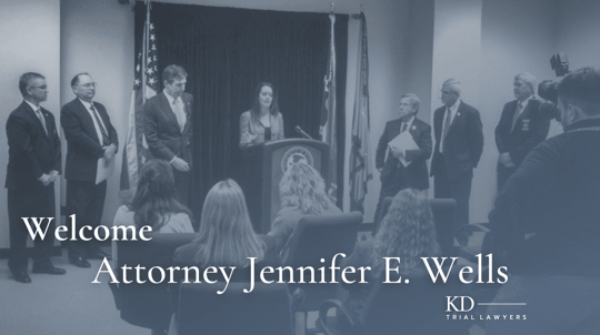 The image shows a formal event with people on stage, one speaking at a podium, and U.S. flags in the background. It welcomes Attorney Jennifer E. Wells to KD Trial Lawyers.