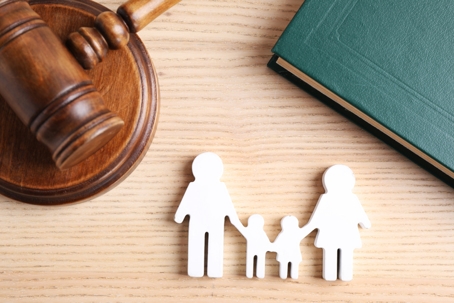 a wooden judge's gavel, a green book, and cut-out figures of a family (two adults and one child). They are placed on a light wooden surface, with the gavel on the left, the book in the upper right, and the family figures in the lower center.