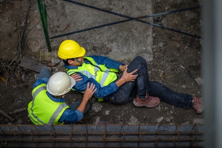 a construction worker lying on the ground, appearing injured, while another worker kneels beside them, providing assistance.