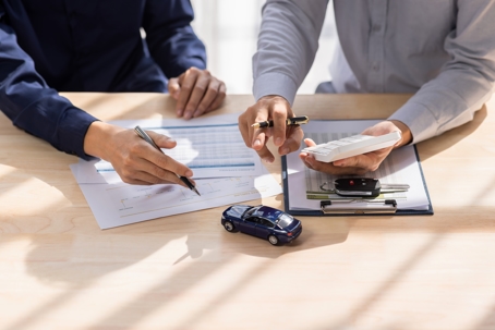 Two people at a table discussing paperwork, with a toy car suggesting a car-related topic, like purchase or insurance.