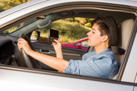 a person sitting in the driver's seat of a car, holding a smartphone in one hand while their other hand rests on the steering wheel