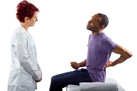 A healthcare professional is engaged in a consultation or examination with a person seated on an examination table. The seated individual is holding their lower back with one hand, possibly indicating discomfort or pain