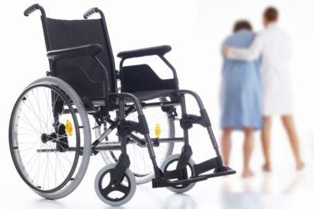 A black wheelchair is positioned in the foreground, showcasing its large rear wheels and smaller front wheels. In the background, a healthcare professional is assisting a patient as they walk, highlighting the support provided to individuals requiring mobility aids.