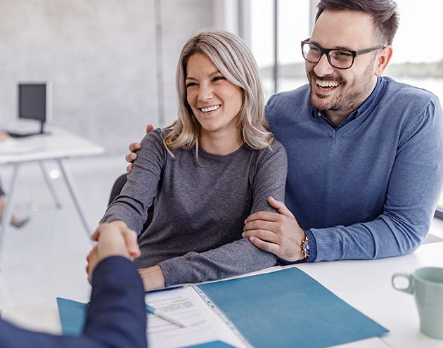 The image shows a professional interaction where one person, standing, is extending their hand in a handshake gesture towards two seated individuals who appear to be a couple.