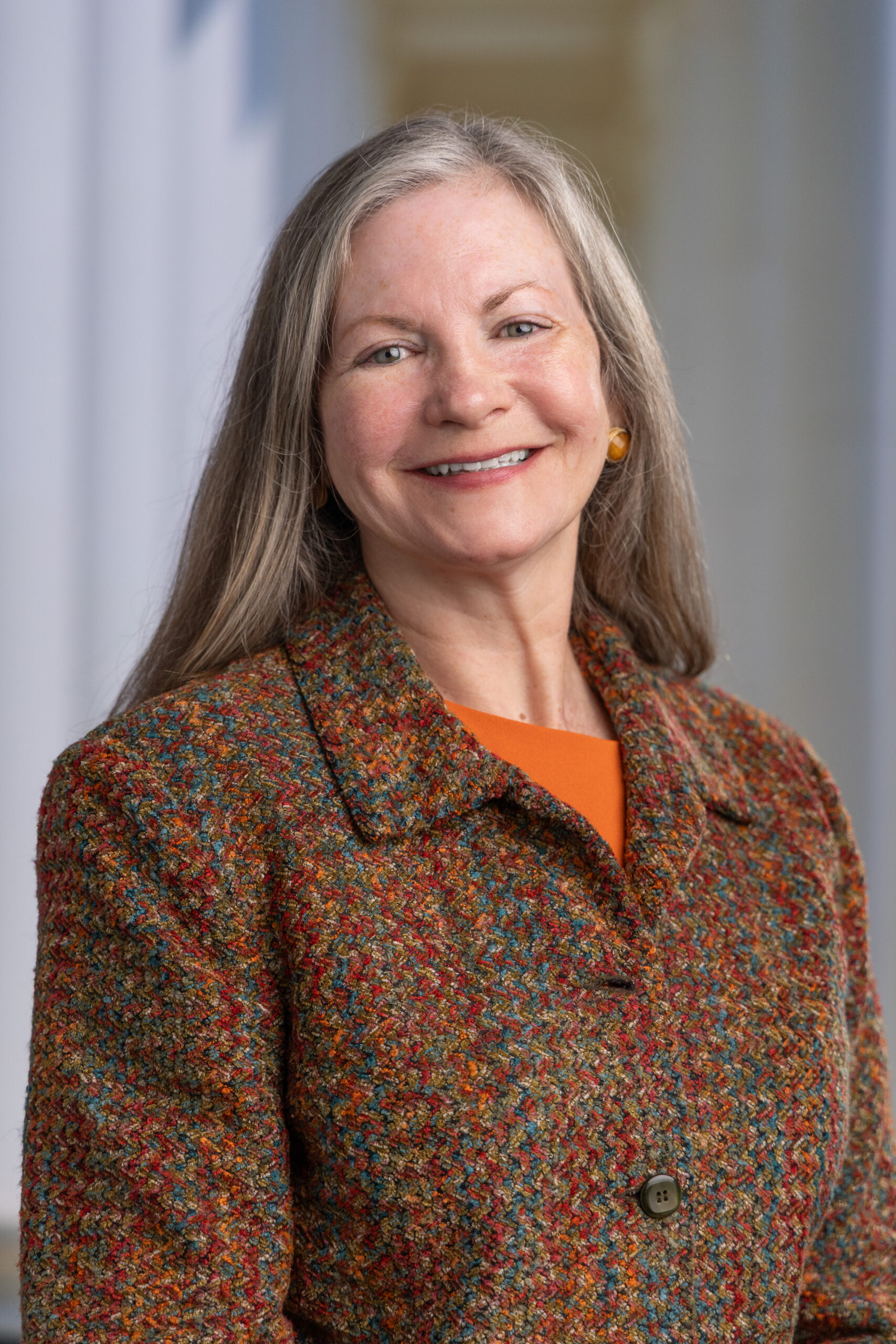 Woman with long gray hair smiles, wearing a colorful patterned jacket over an orange top, in a bright indoor setting.