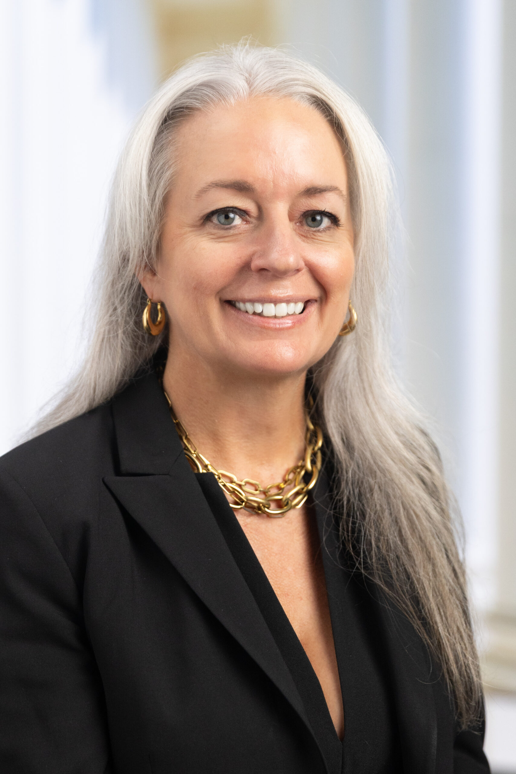 Woman with long gray hair, wearing a black blazer and gold chain necklace, smiling against a light background.