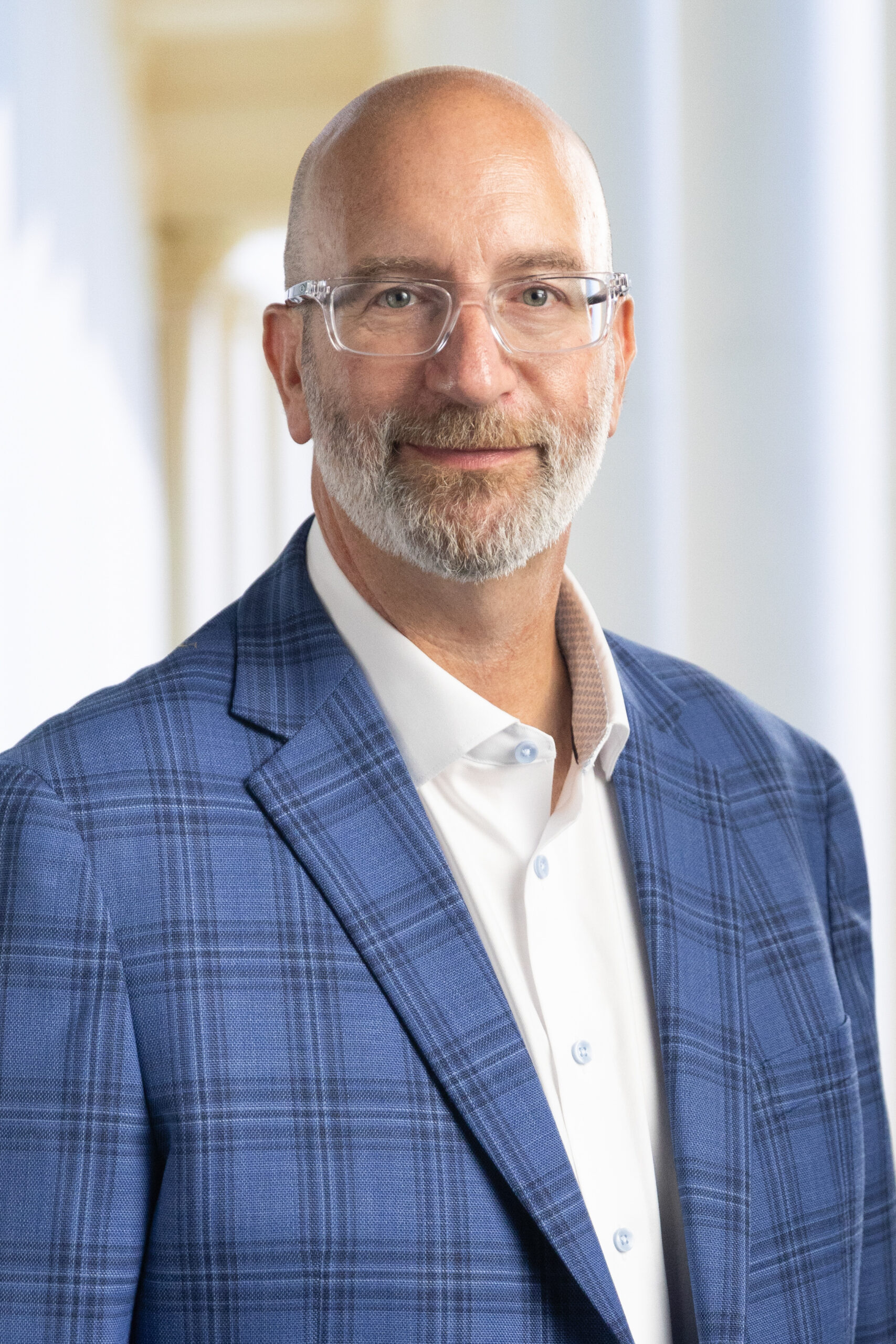 Man with glasses and a beard wearing a blue plaid blazer over a white shirt, smiling in a bright corridor.