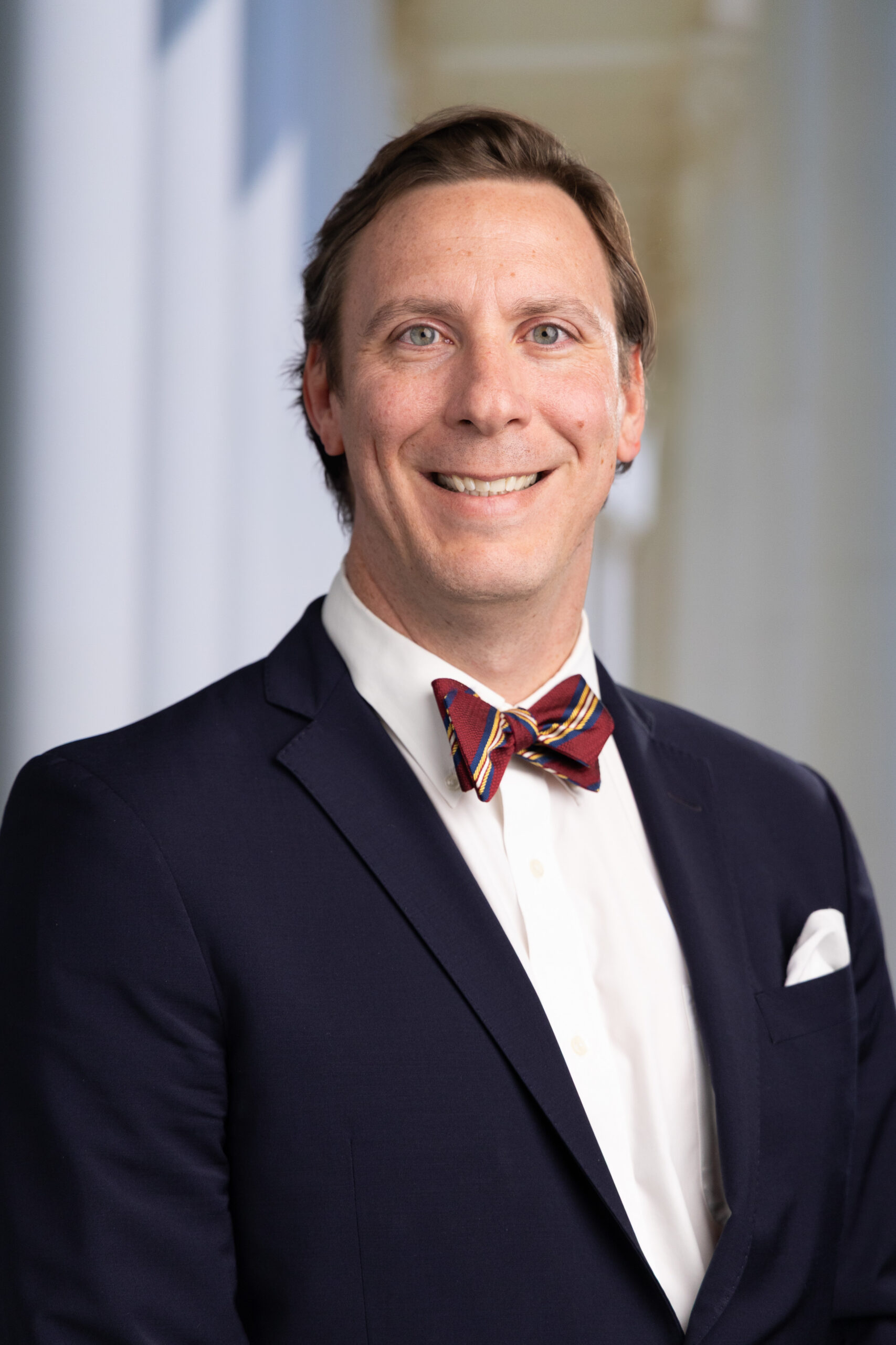 Man in a navy suit and white shirt, smiling, wearing a colorful striped bow tie, with a blurred background.