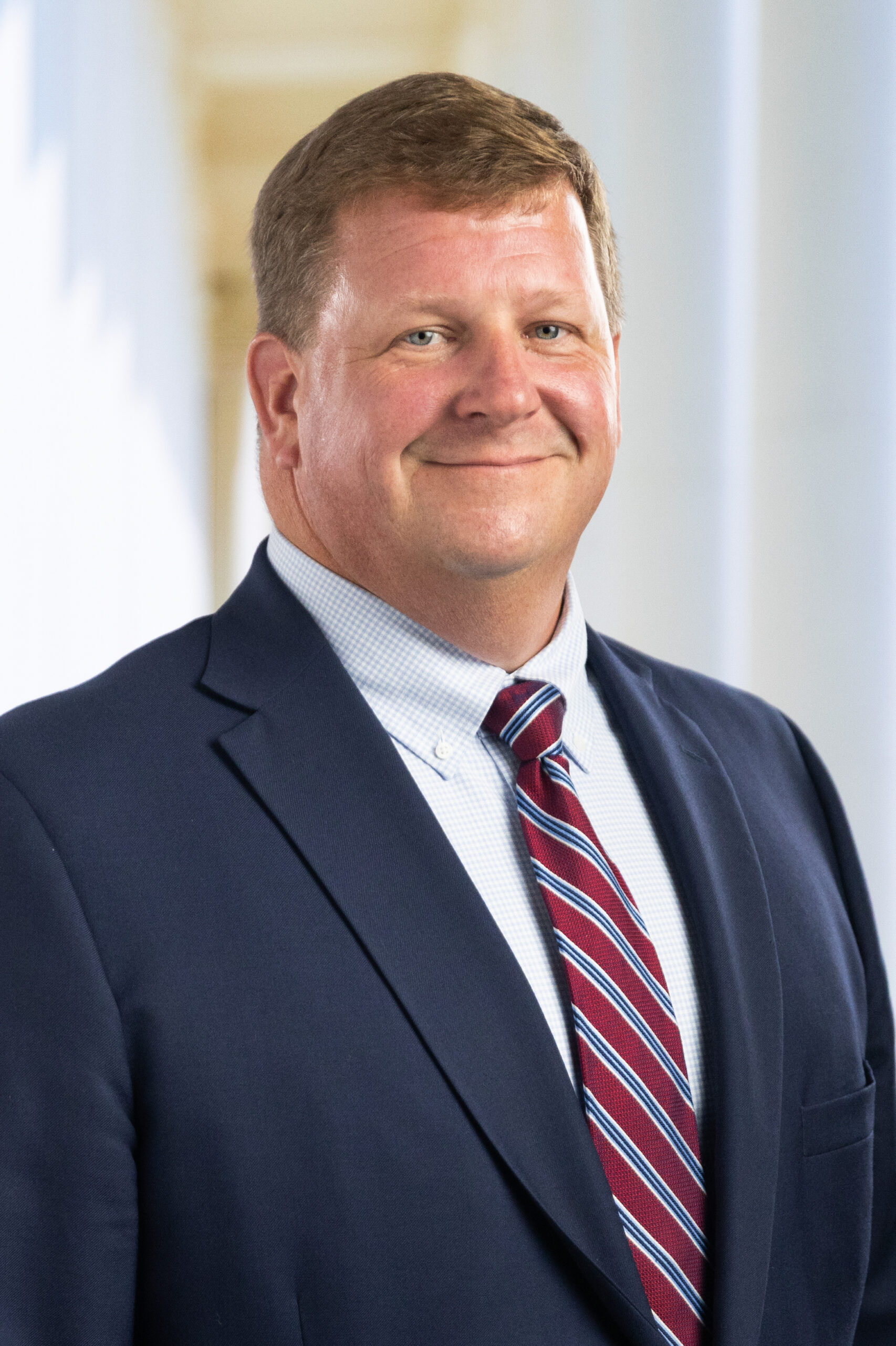 Smiling man in a navy suit and striped red tie, standing in a bright, columned setting.