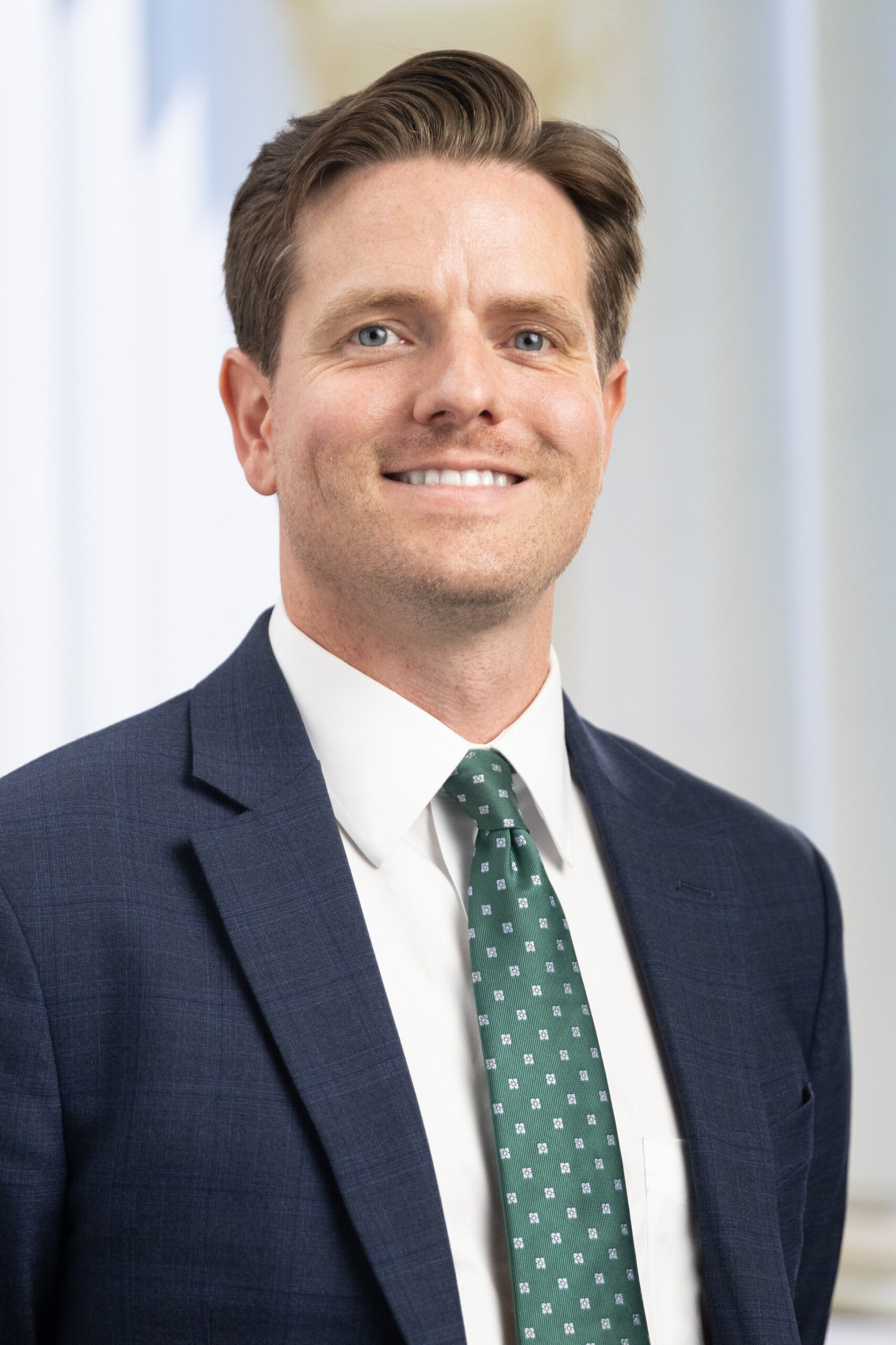 Man smiling in a navy suit and white shirt, wearing a green patterned tie, with a light background.