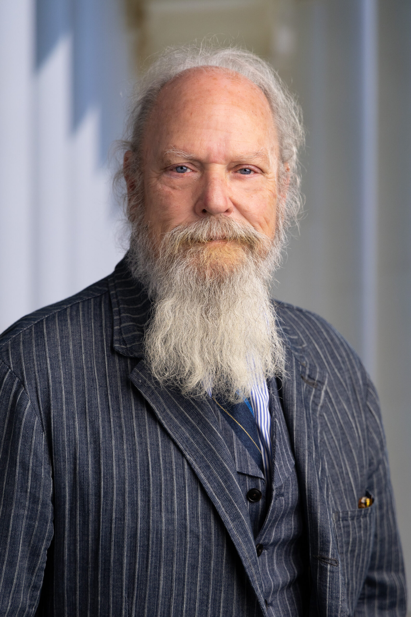 Older man with a long white beard, wearing a pinstriped suit, standing against a light background.