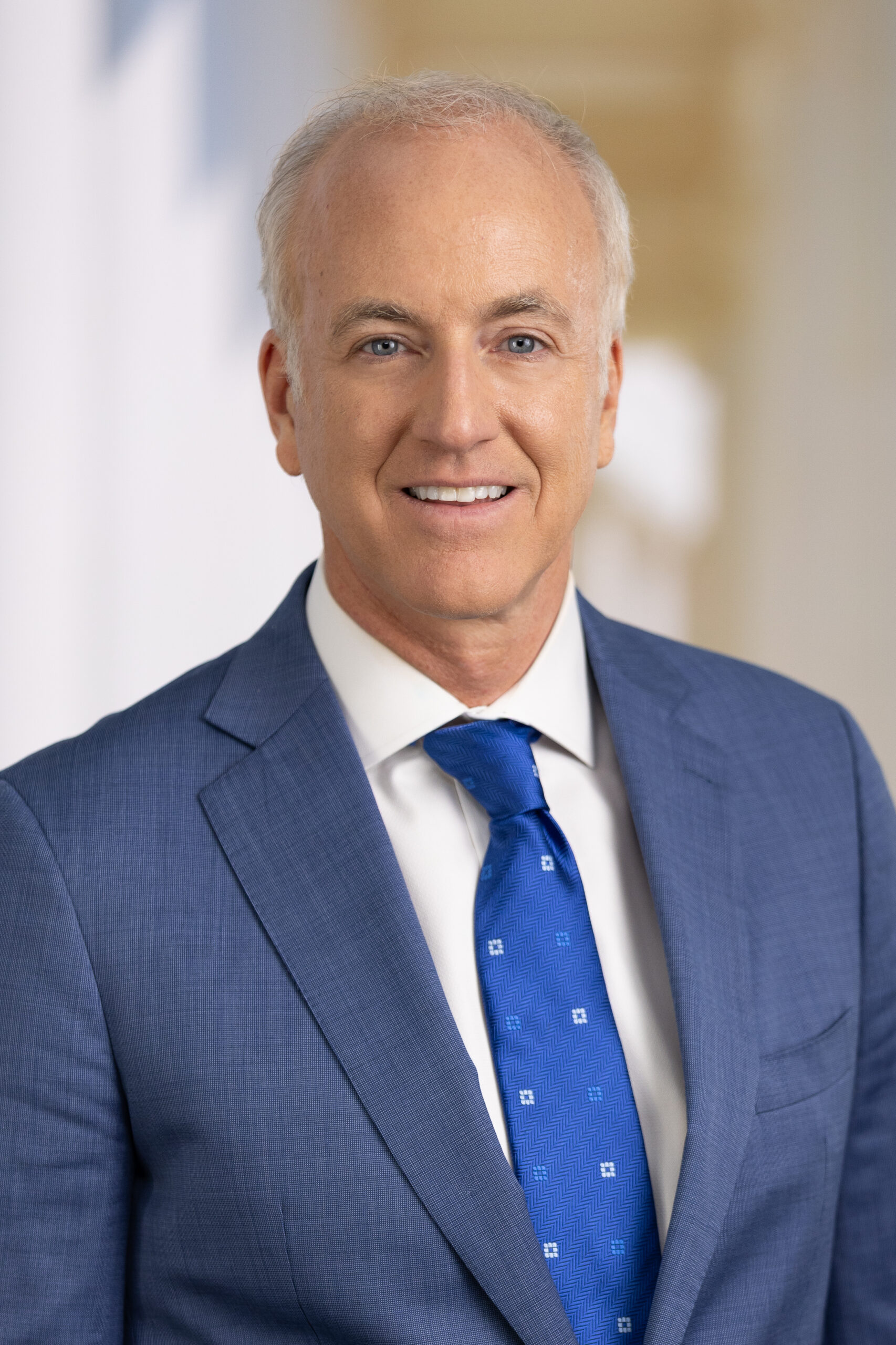 Man in a blue suit and patterned tie smiles against a blurred background.