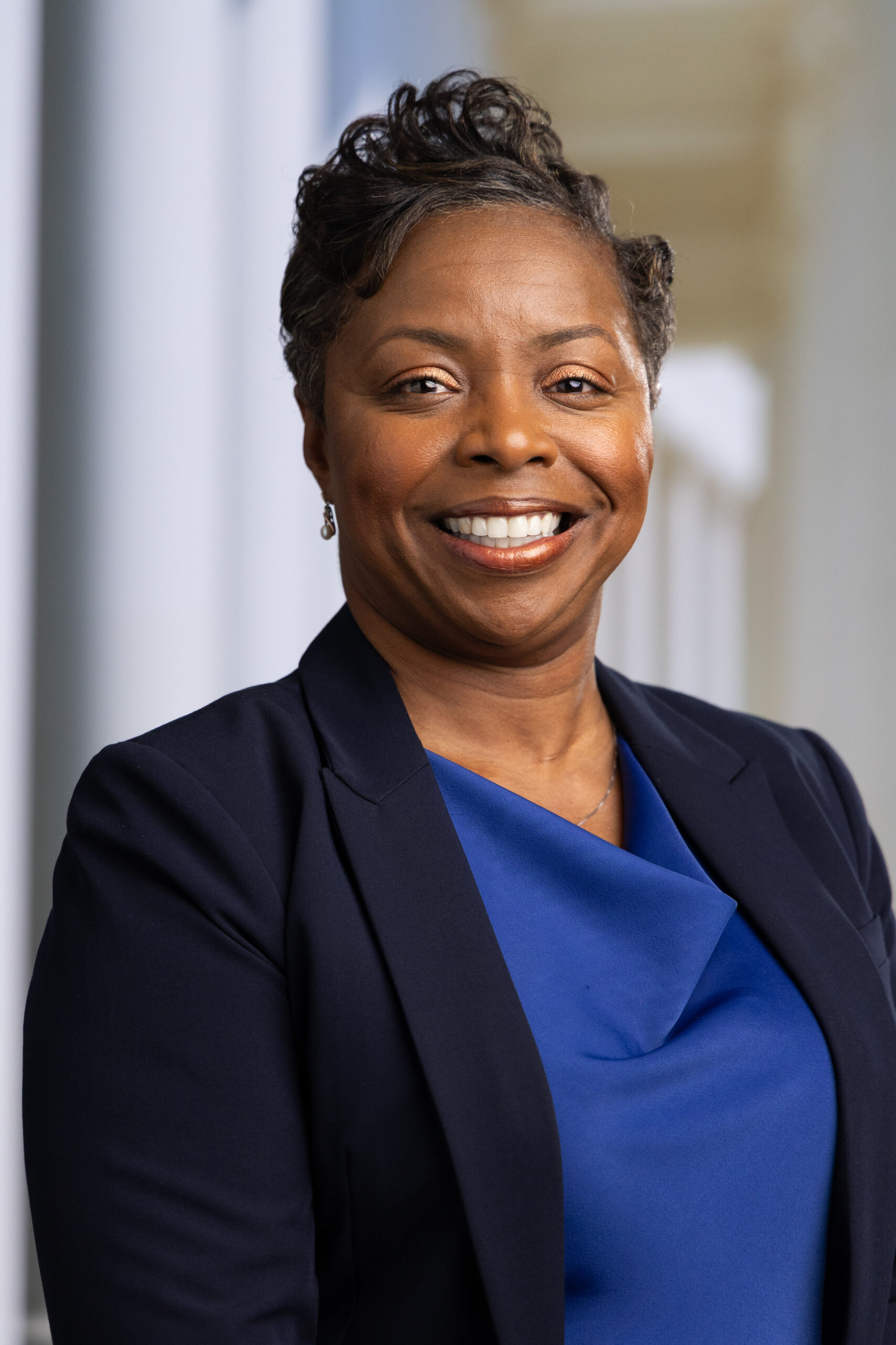 Smiling woman in a navy blazer and blue top, standing in a bright, modern setting.