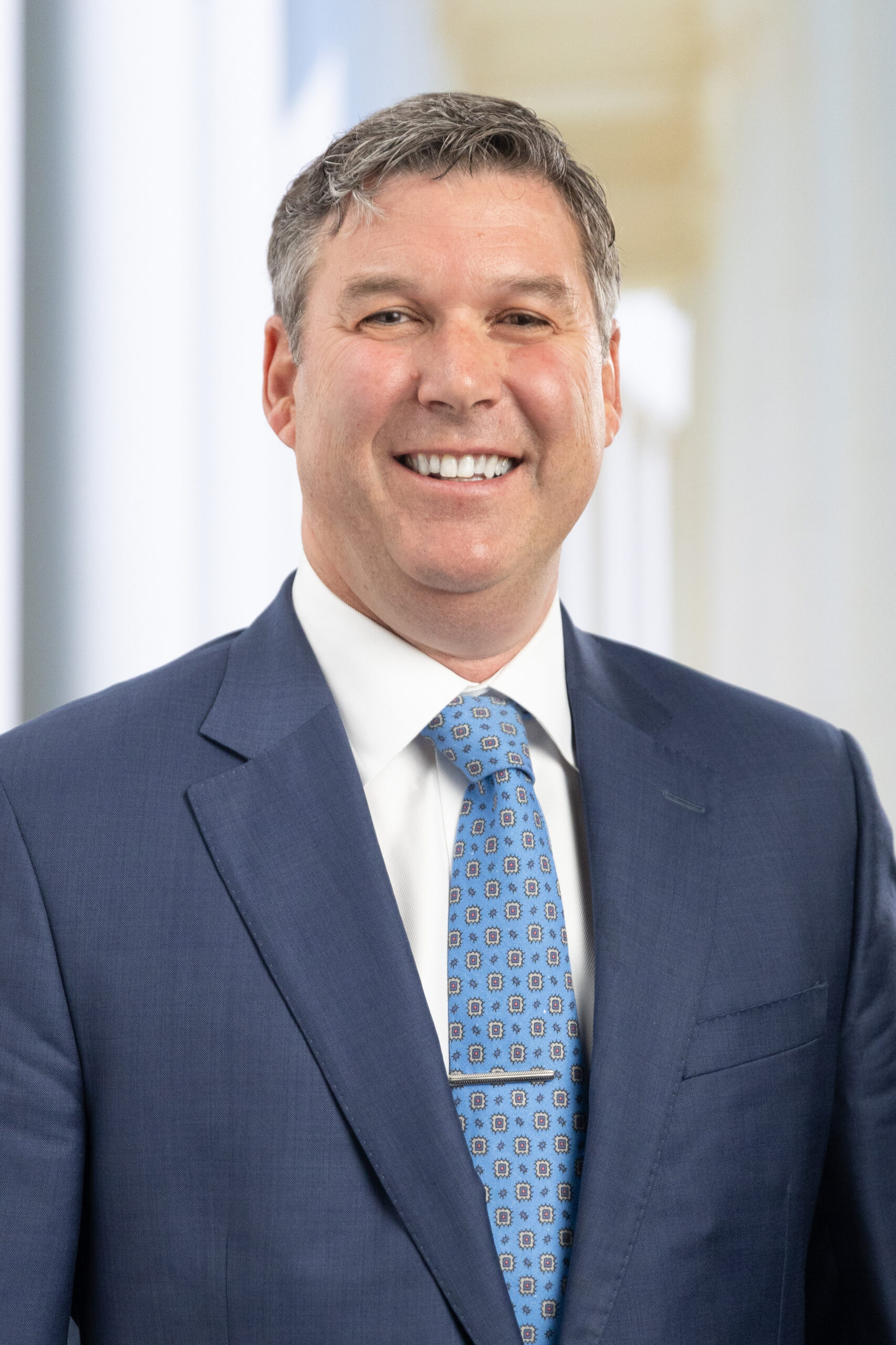 Smiling man in a blue suit and patterned tie, standing in a bright, modern interior.