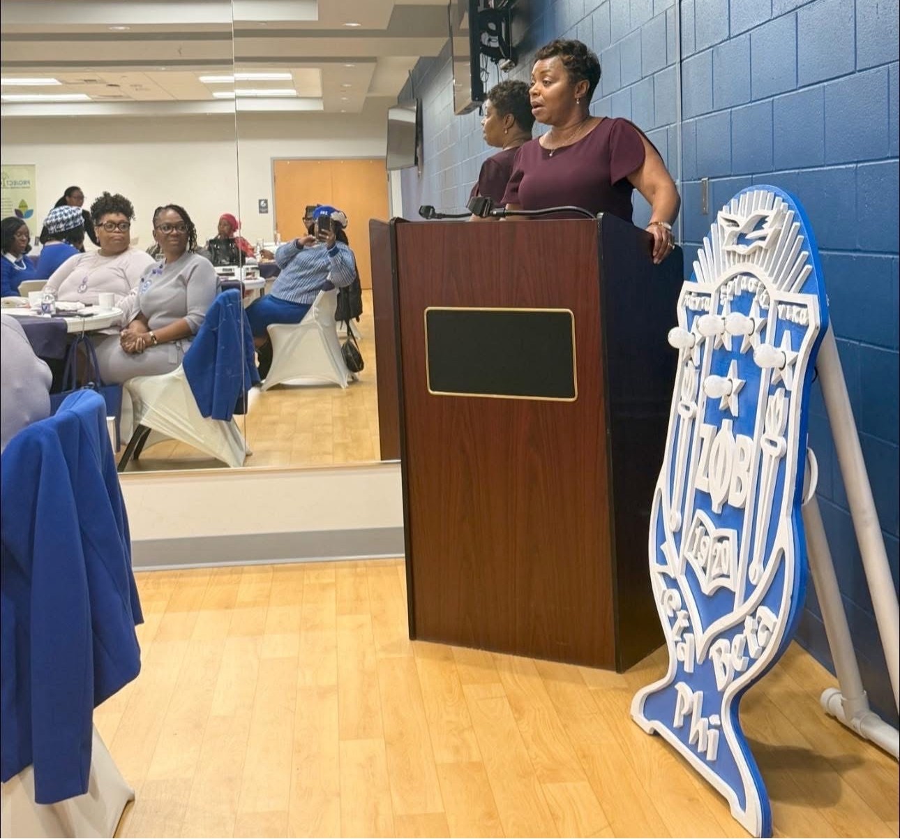Woman speaking at a podium in a blue and white decorated room, with attendees seated and a logo displayed nearby.