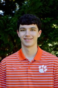 Young man with dark curly hair wearing an orange and white striped polo shirt with a Clemson logo, smiling outdoors.